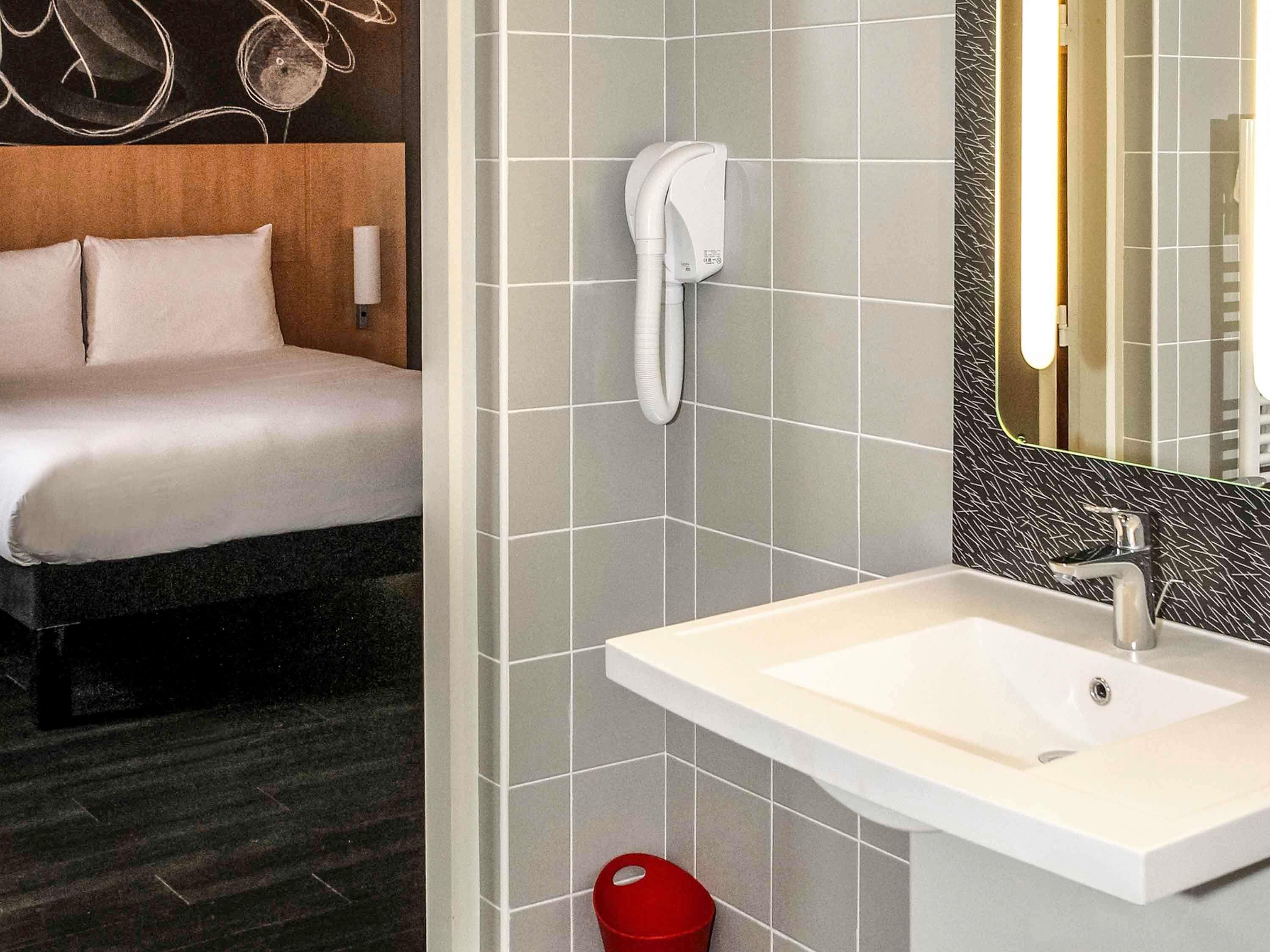 A hotel room with a bed, a bathroom with a sink and mirror, and a red trash bin.