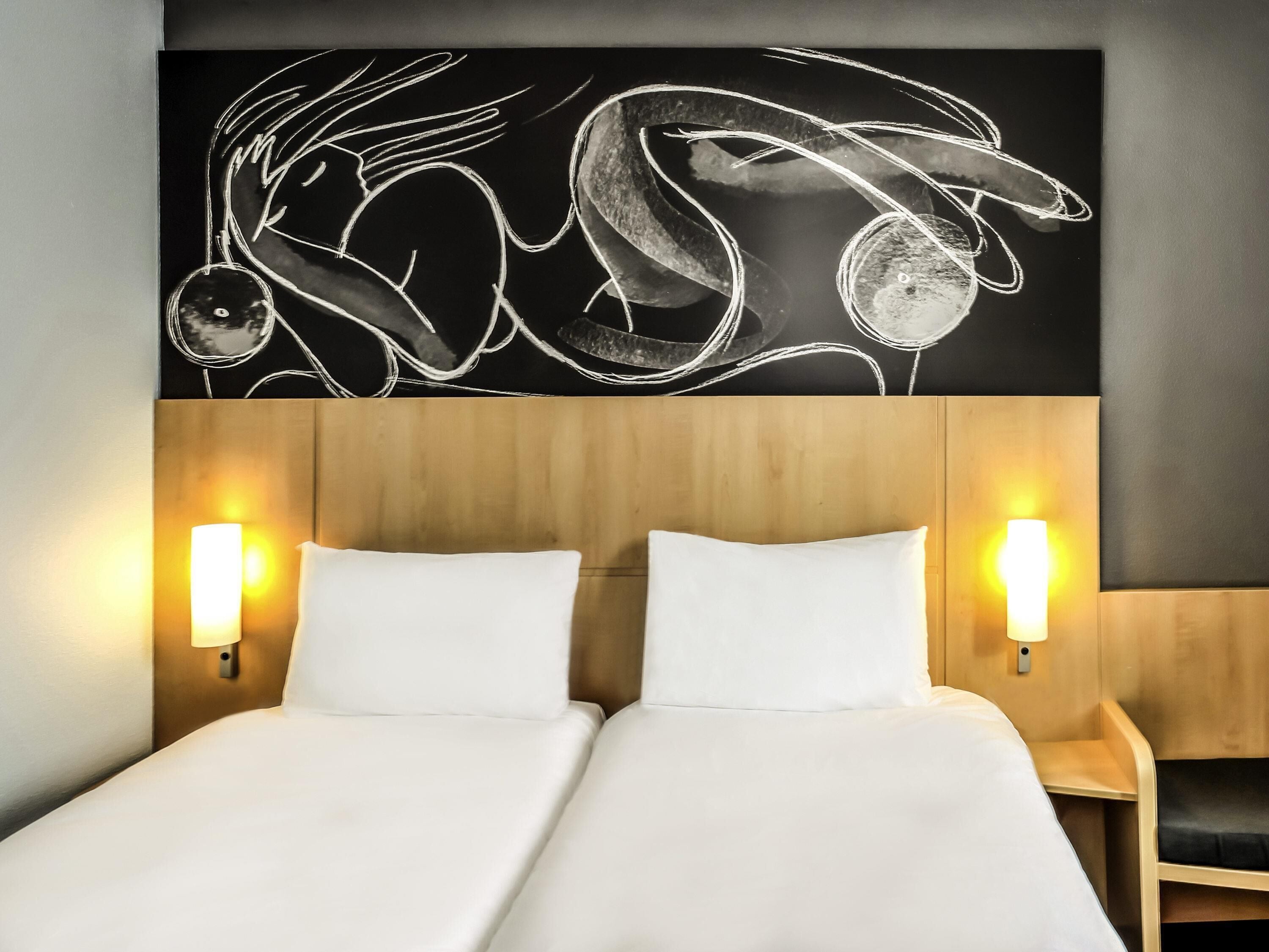 Een tweepersoonsbed met witte lakens, twee wandlampen langs het bed en een groot abstract kunstwerk erboven.