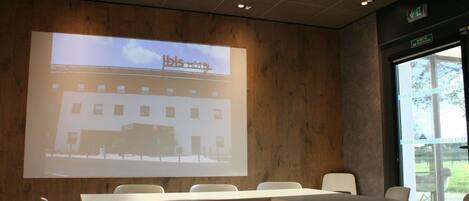 Sala de conferências com tela de projeção exibindo uma imagem de hotel, mesas e cadeiras brancas.