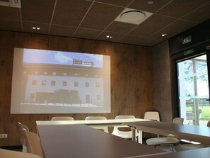 A conference room with a projection screen displaying a hotel image, white tables, and chairs. - Ibis Montceau Les Mines (Montceau-les-Mines)