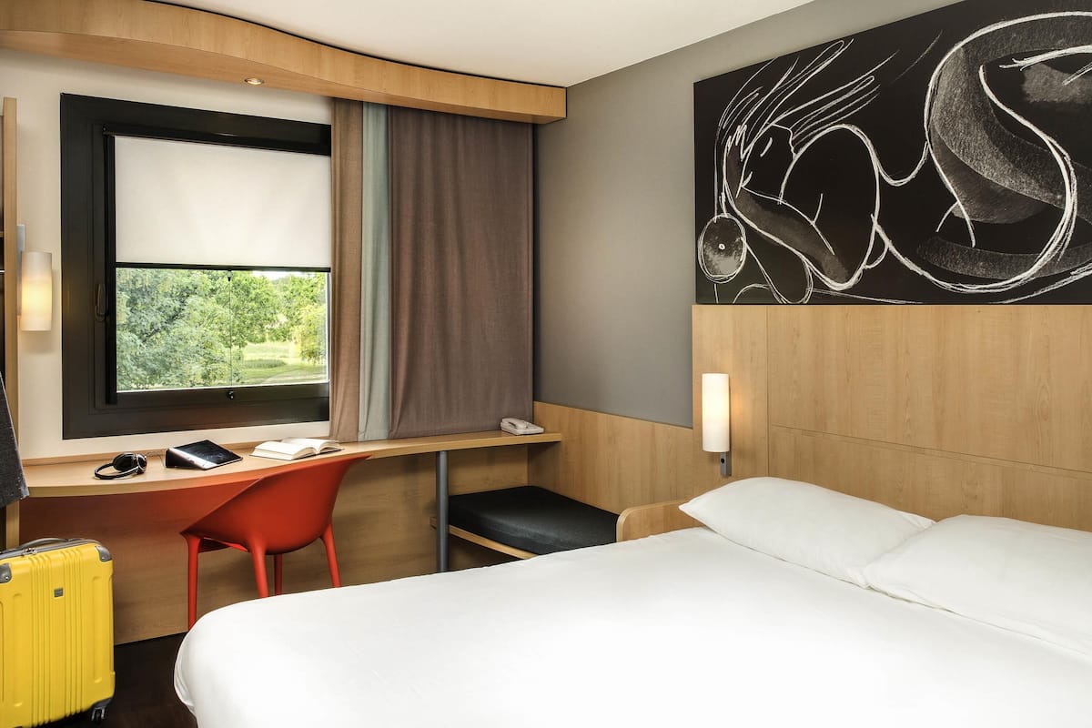 A hotel room with a bed, a desk with a chair, a window with curtains, a wall-mounted phone, and a yellow suitcase.