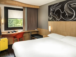 A hotel room with a bed, a desk with a chair, a window with curtains, a wall-mounted phone, and a yellow suitcase.