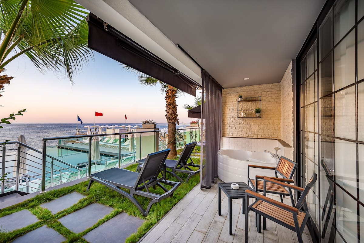 A balcony with a view of the sea, lounge chairs, a table, and a bathtub.