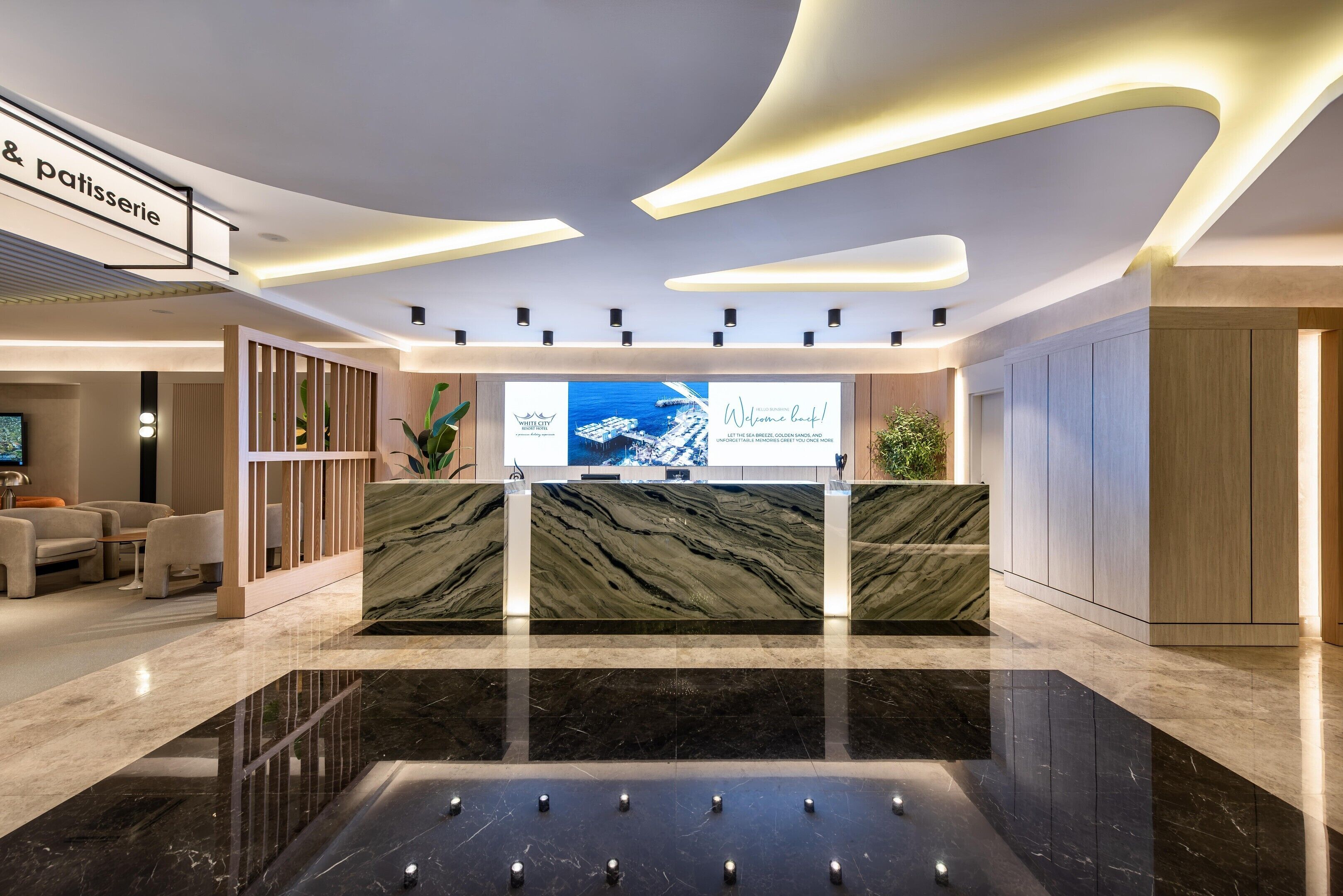 A modern hotel lobby with a reception desk, seating area, and a large screen displaying the hotel's name.