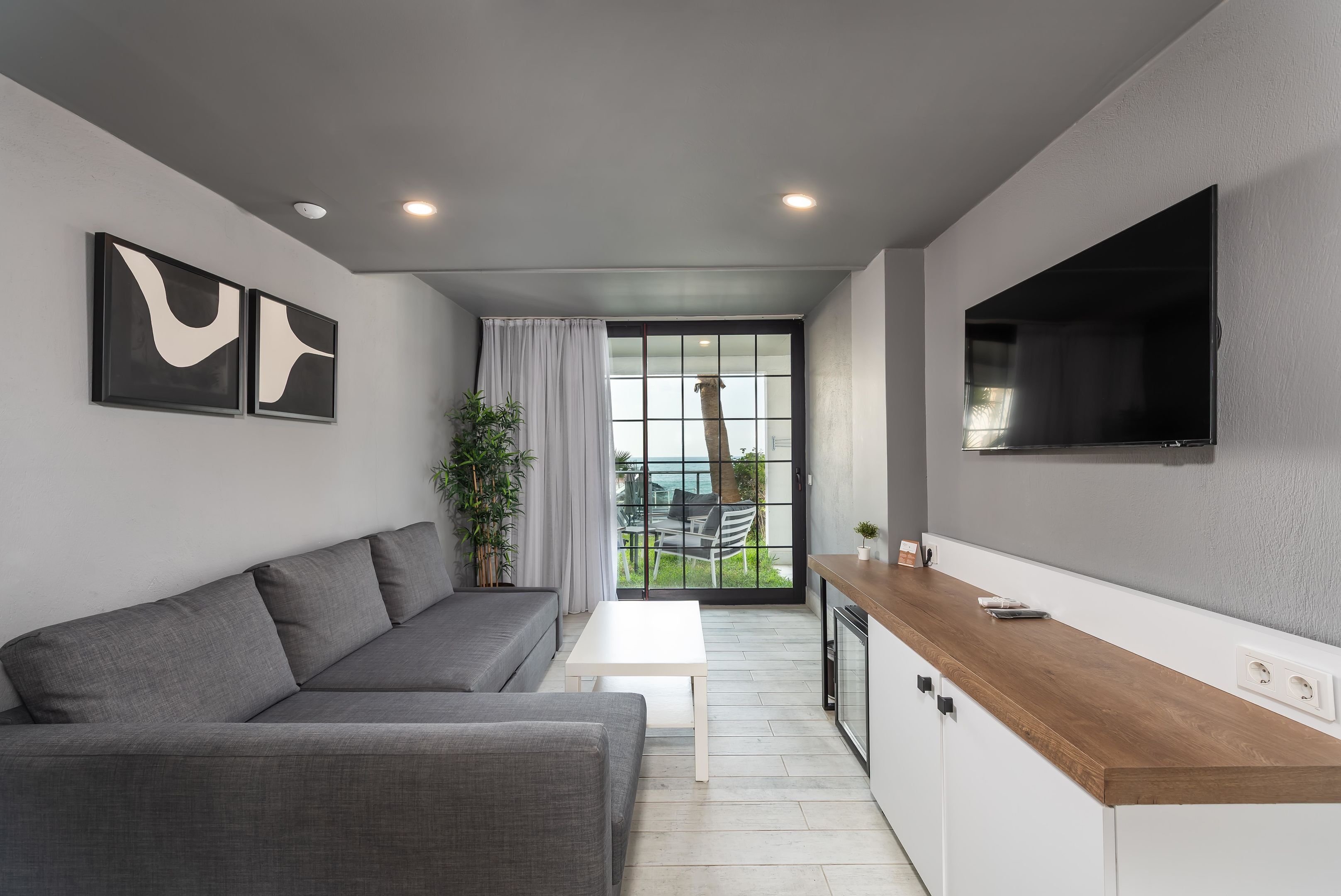 Un espacio de estar moderno con un sofá gris, una mesa blanca, una encimera de madera y un televisor instalado en la pared.