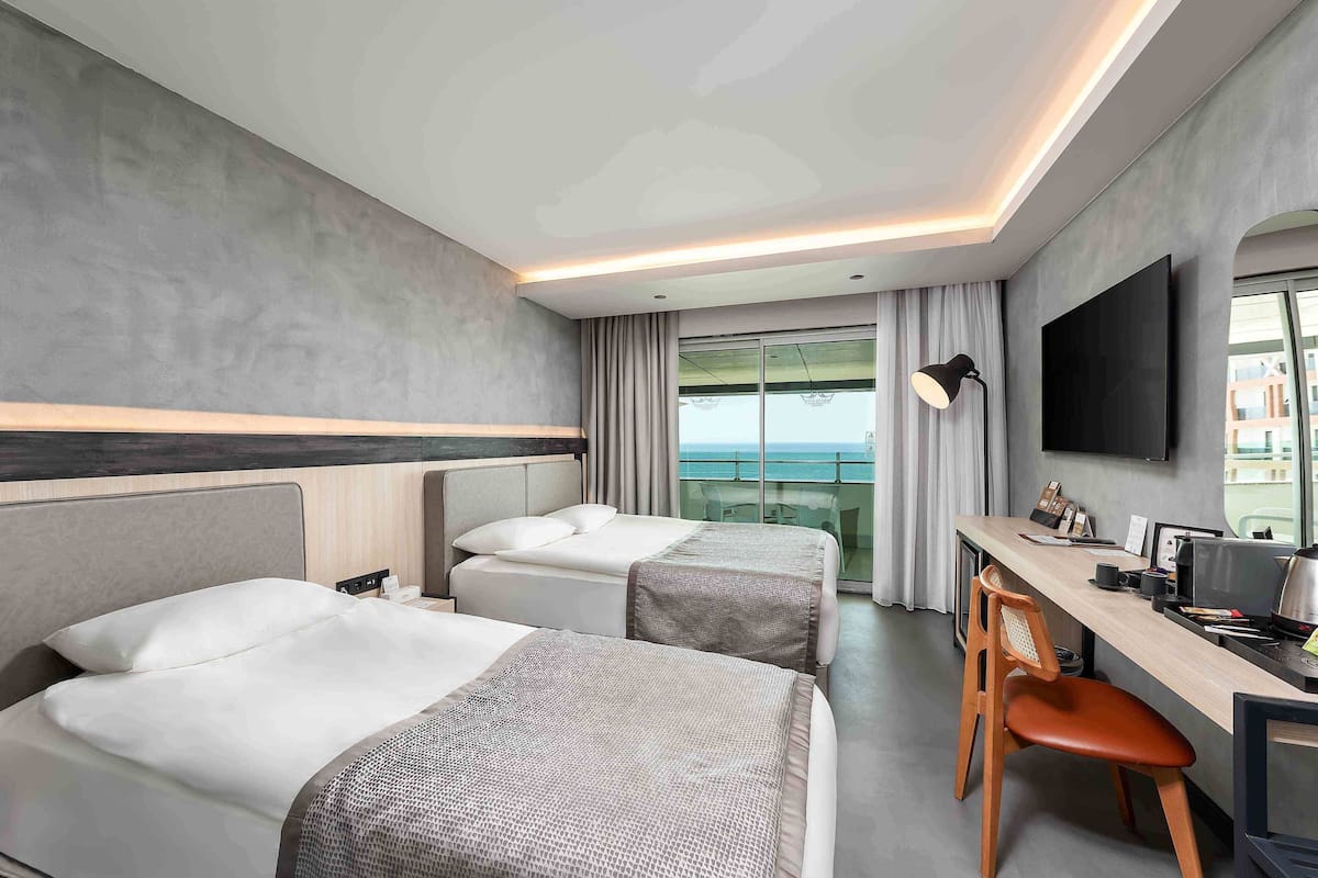 A hotel room with two beds, a desk, a chair, a TV, and a balcony with a view.