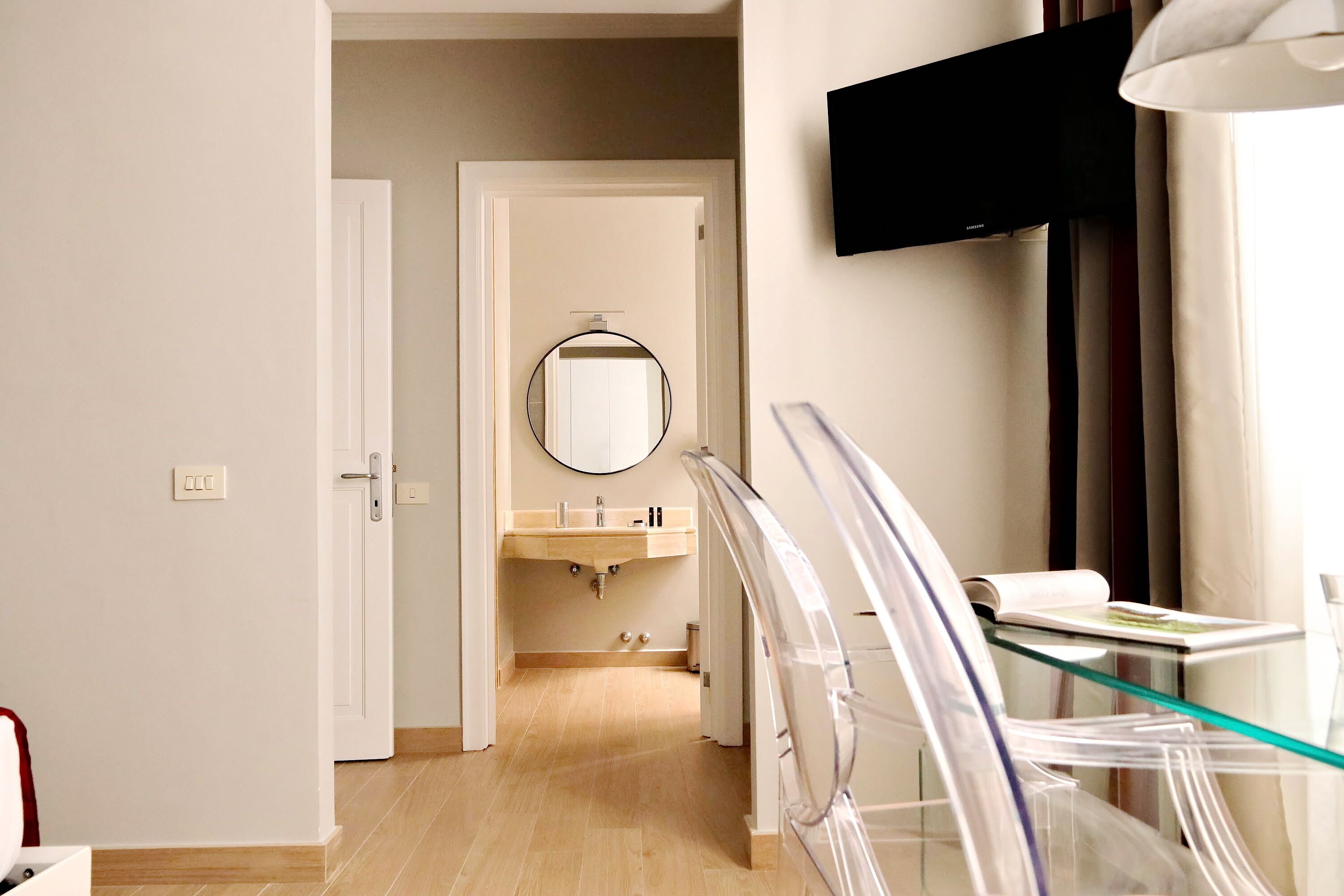 Une chambre d’hôtel avec un plancher en bois, une table en verre, un téléviseur fixé au mur et une salle de bain équipée d’un lavabo et d’un miroir.