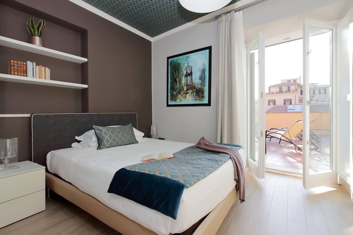 A modern hotel room with a large bed, a bookshelf, a painting, and a balcony with a view of buildings.