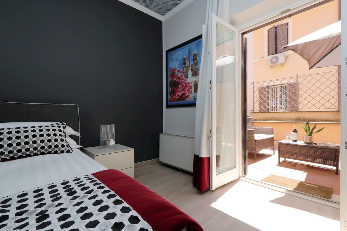A bedroom with a bed, a nightstand, a glass of water, a red blanket, a painting, and a balcony with a wicker table and chairs.