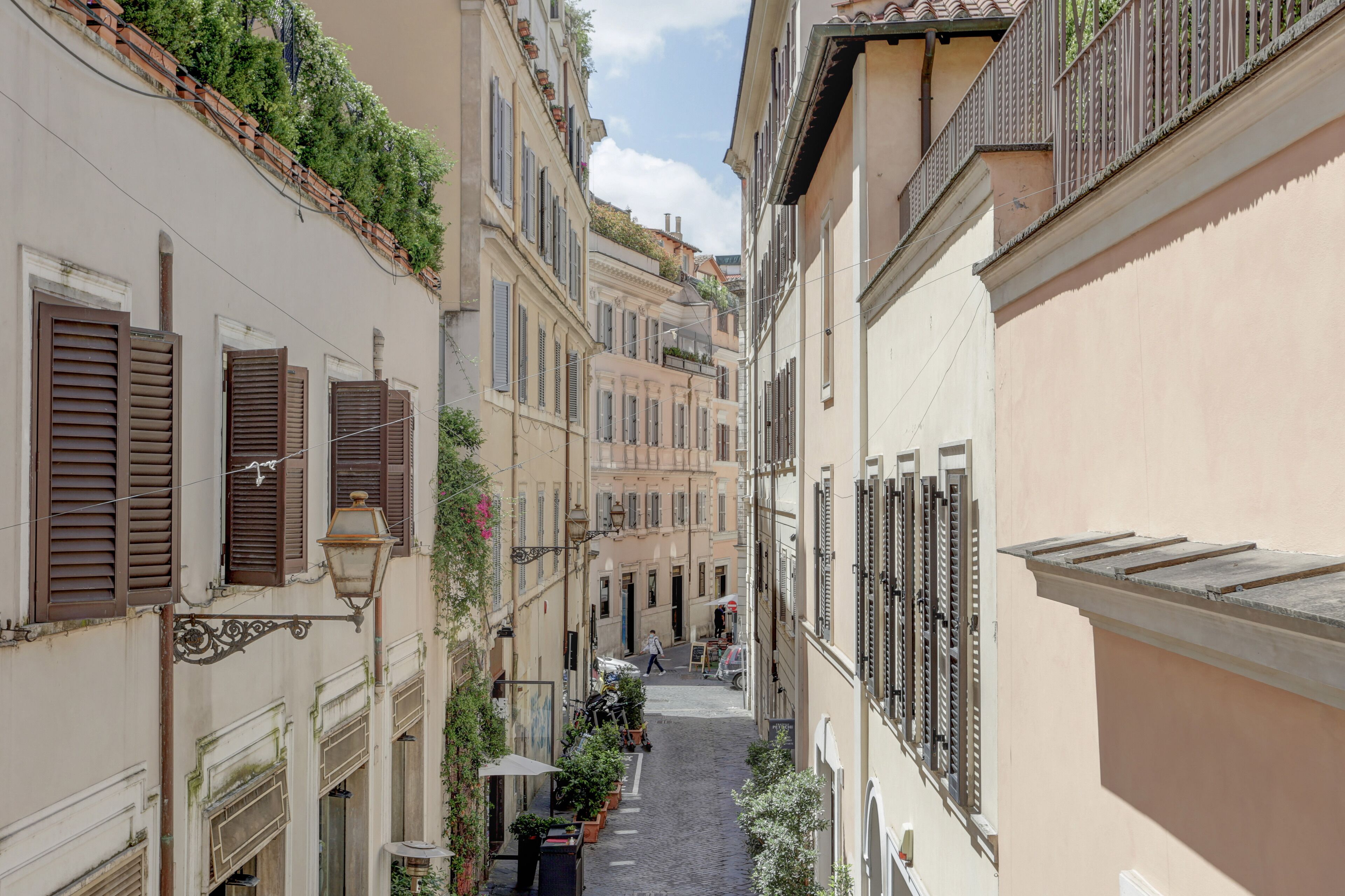 A narrow street flanked by buildings with shutters and balconies, a cobblestone path, and outdoor seating areas.