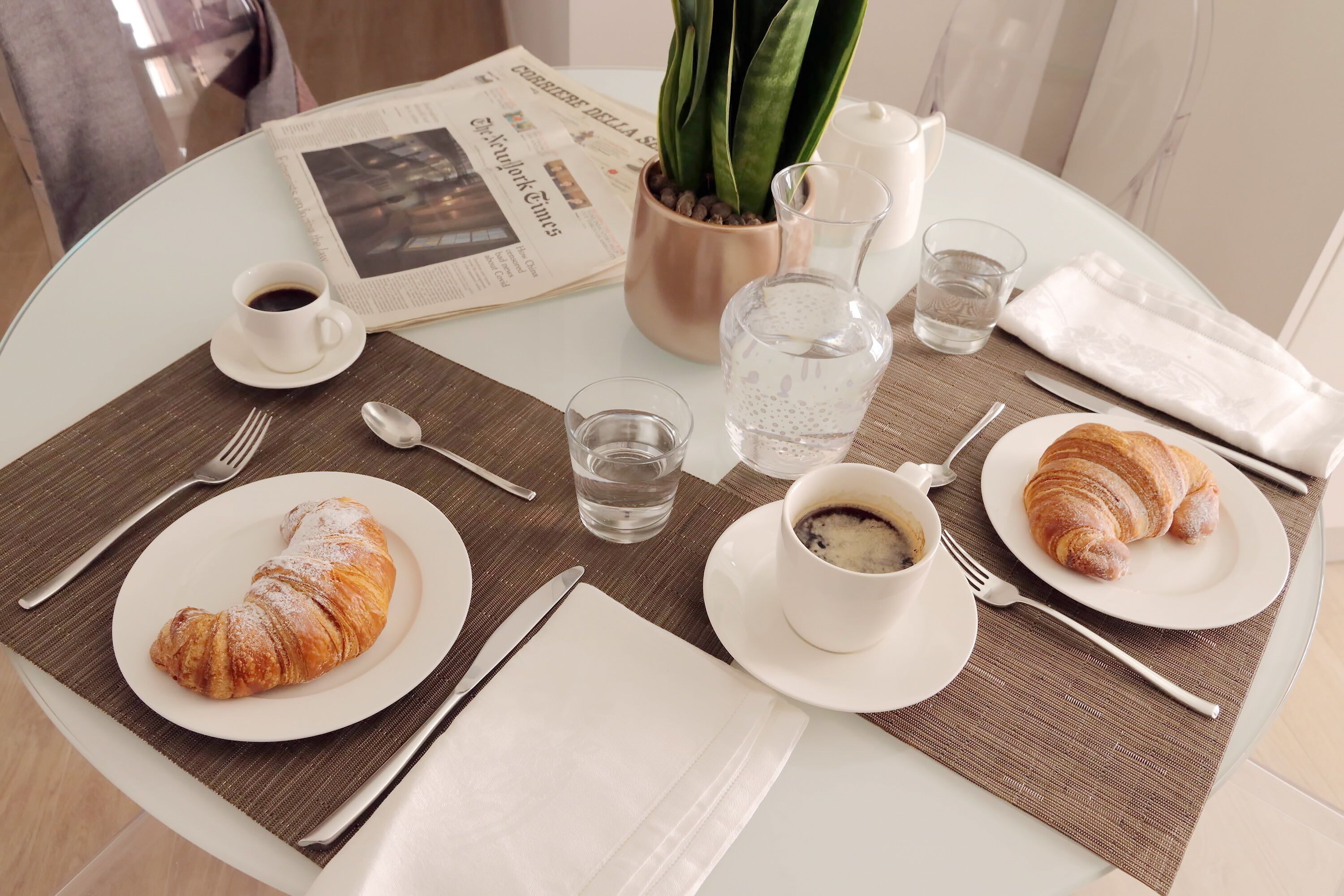 A breakfast setting with croissants, coffee, and a newspaper on a glass table.