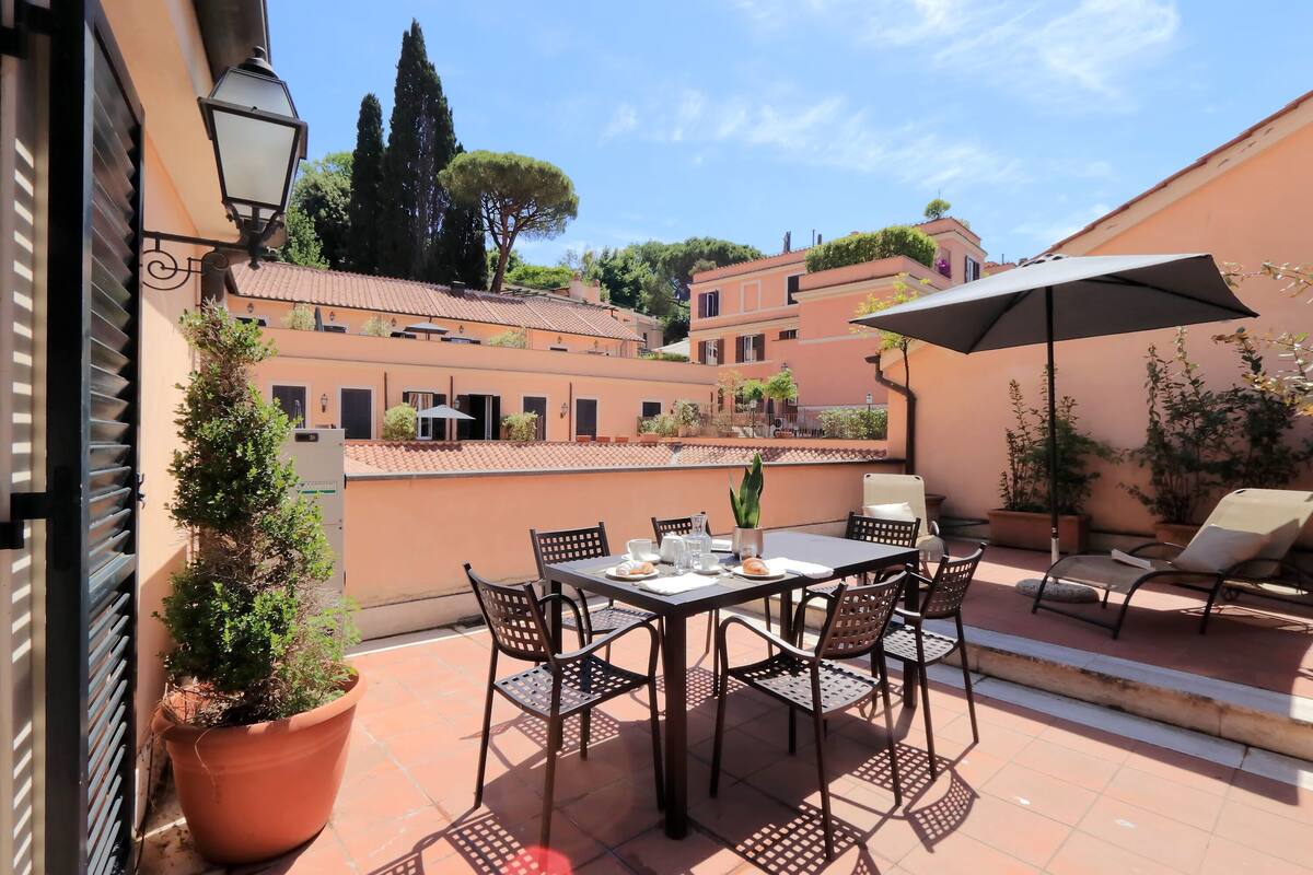 An outdoor patio with a table and chairs, surrounded by potted plants and a large umbrella.