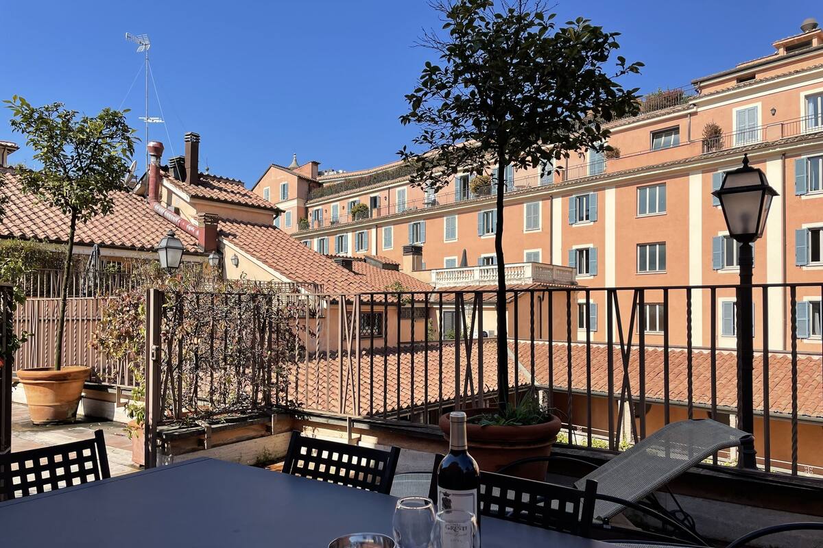 A terrace with a table, wine bottle, and glasses, overlooking a residential area with terracotta roofs and balconies.