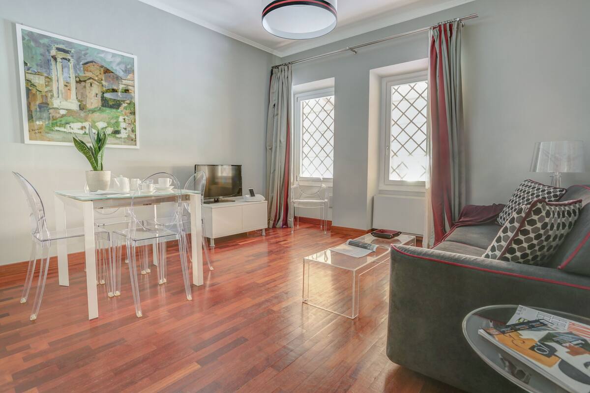 A modern living room with a glass dining table, clear chairs, a grey sofa, and a television.