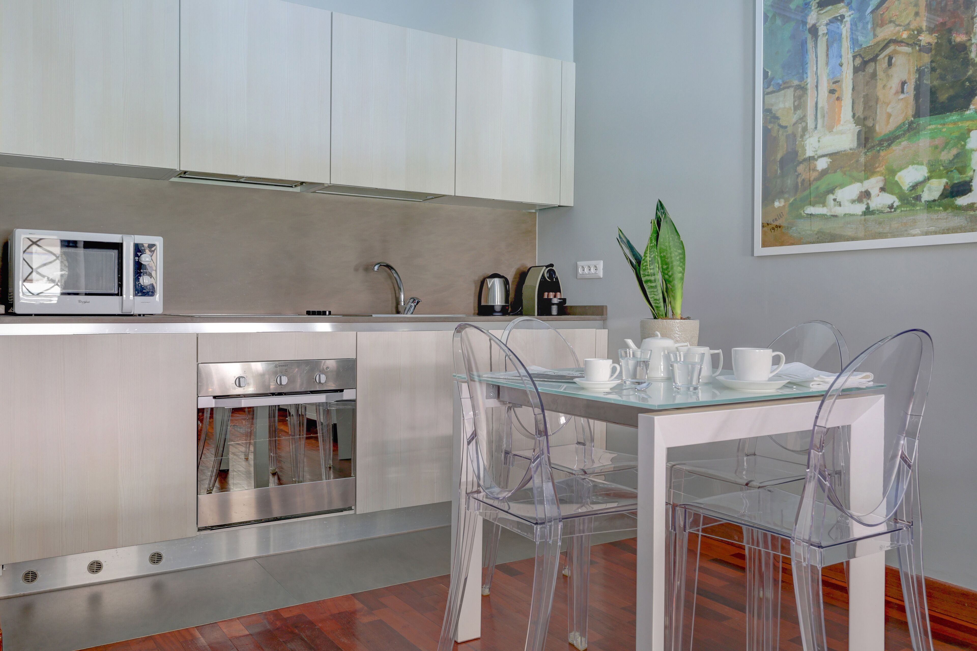 A modern kitchen with white cabinets, stainless steel appliances, a glass dining table with clear chairs, and a potted plant.