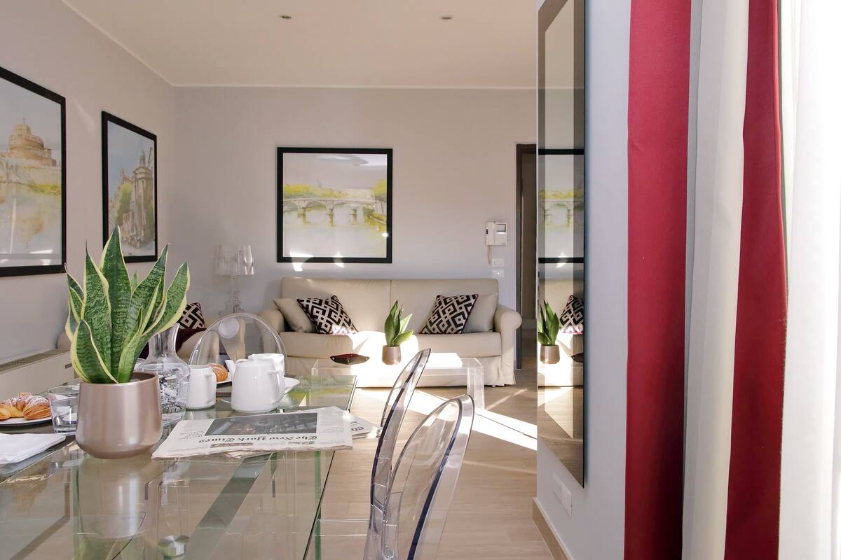 A modern living room with a glass dining table, clear chairs, a sofa, and a potted plant.