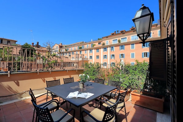 A terrace with a table and chairs, a lamp, and a view of a building with balconies and windows.