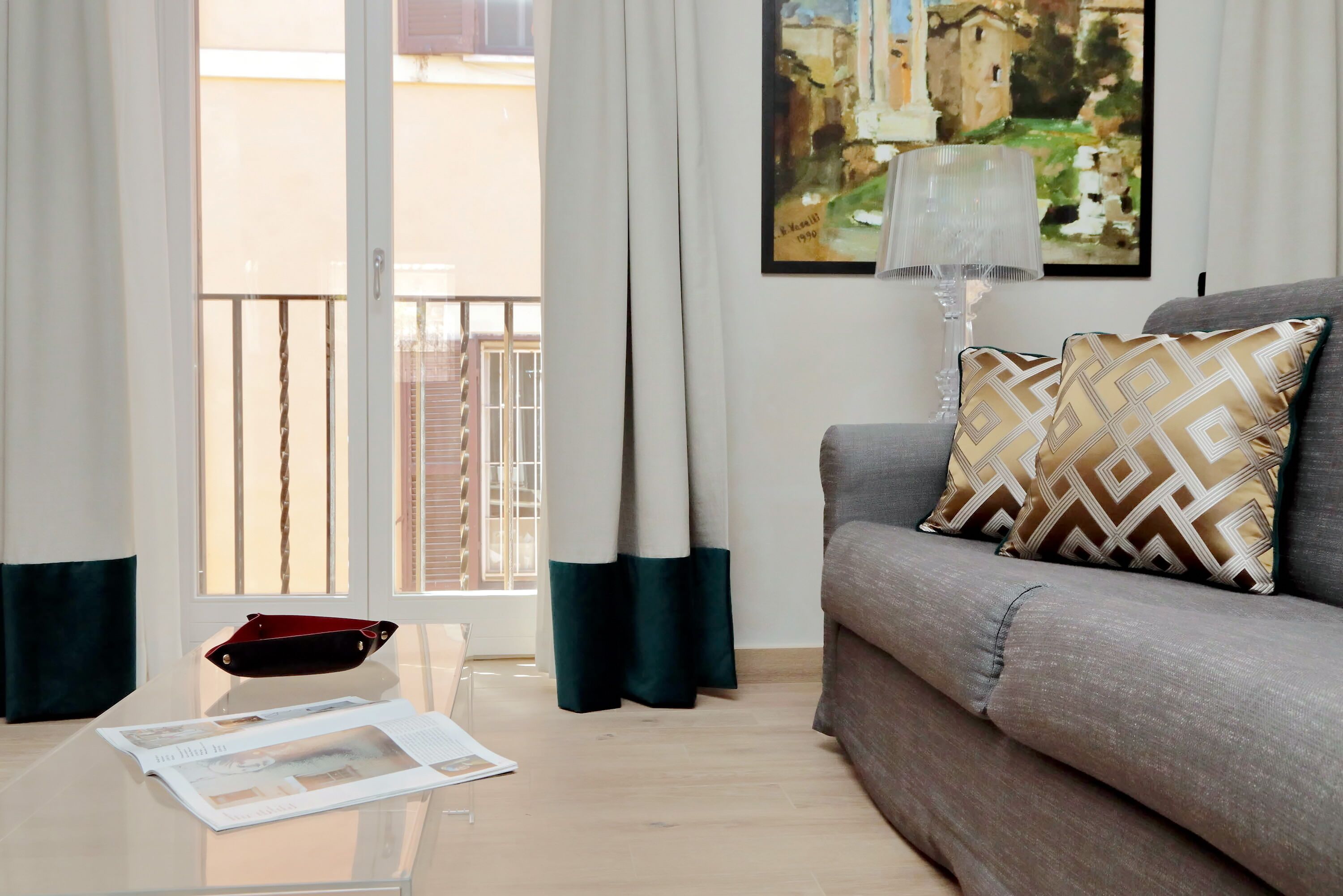 A living room with a sofa, a glass coffee table, a magazine, a lamp, and a painting on the wall.