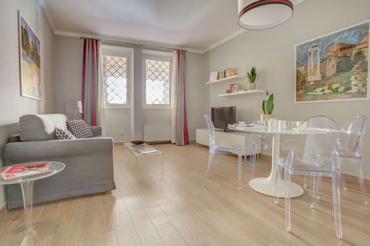 A modern living room with a grey sofa, a glass coffee table, and a dining area with a glass table and clear chairs.