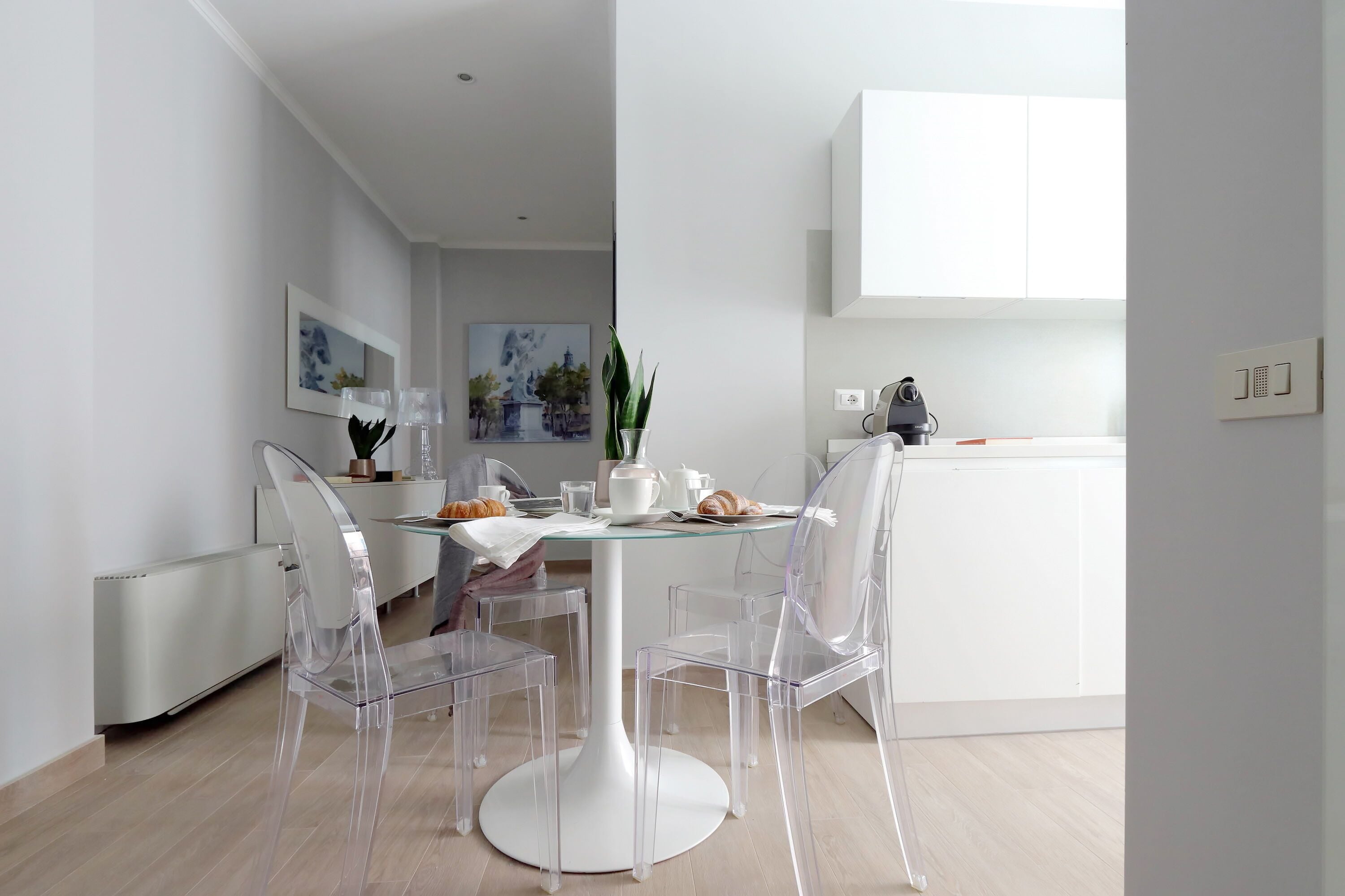 A modern dining area with a glass table and clear chairs, white walls, and minimalistic decor.