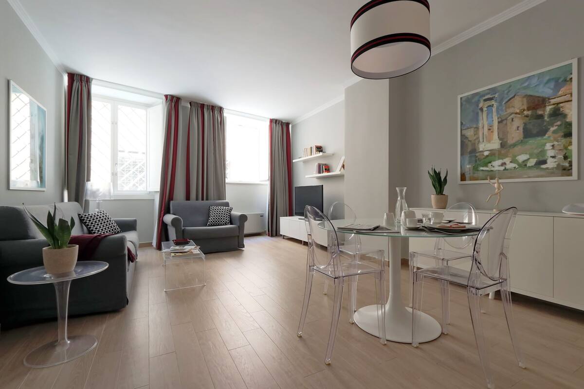 A modern living room with a glass dining table, clear chairs, a grey sofa, and a potted plant.