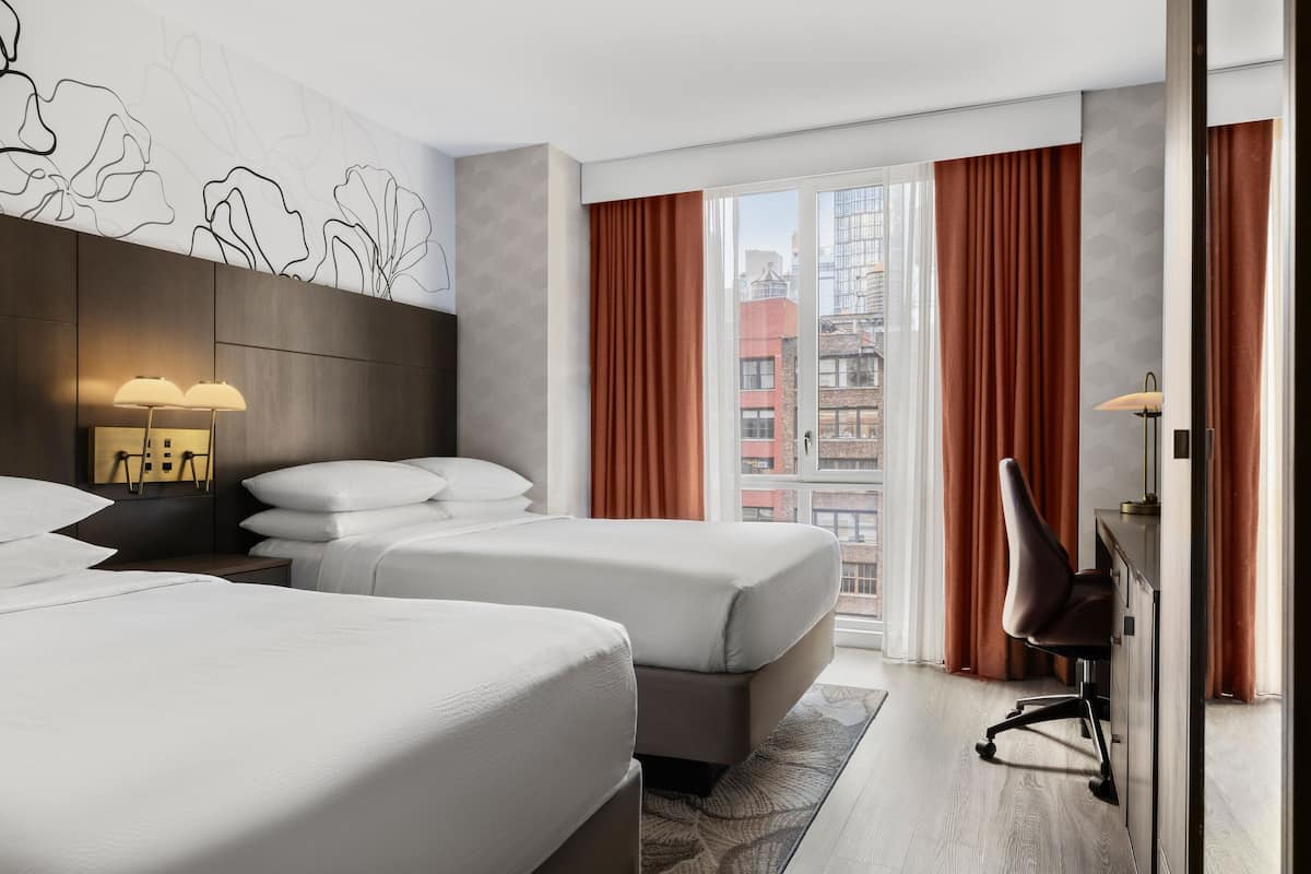 Ein Hotelzimmer mit zwei Betten, einem Schreibtisch, einem Stuhl und einem großen Fenster mit Vorhängen.