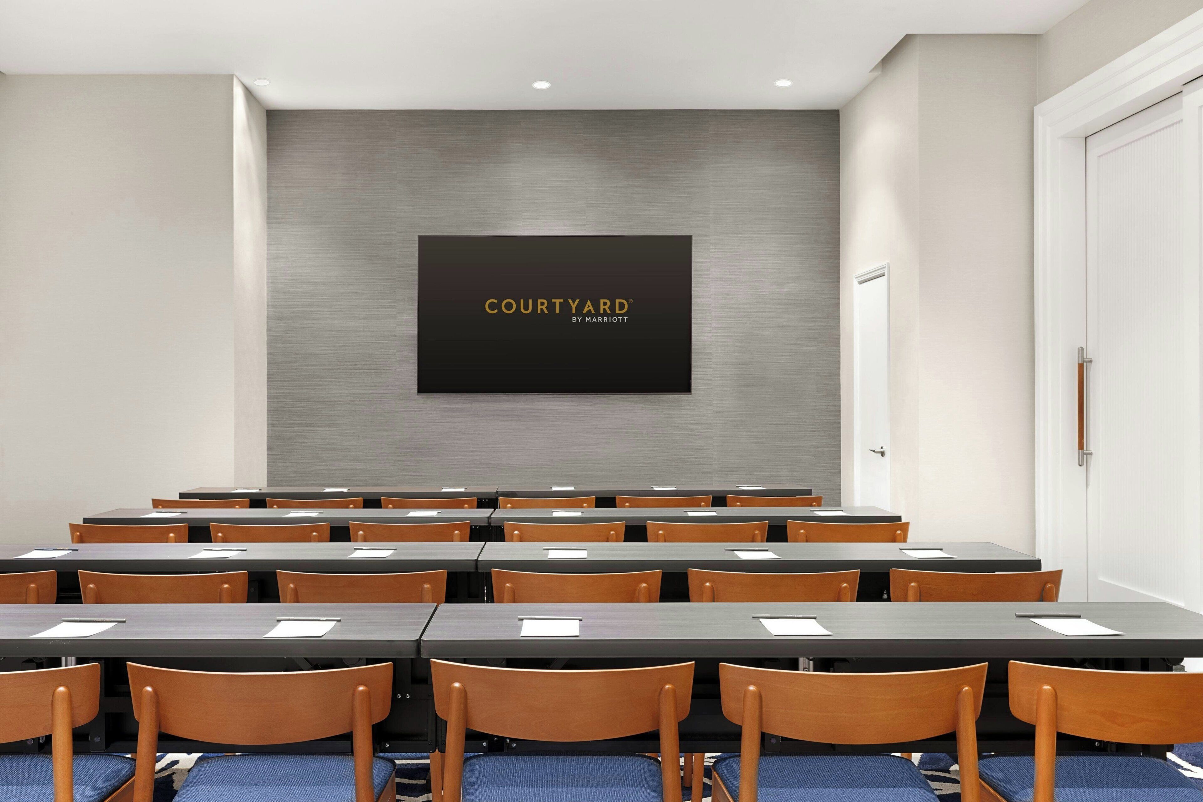 Ein Konferenzraum mit mehreren Tischen und Stühlen, einem großen Bildschirm, der "COURTYARD BY MARRIOTT" anzeigt, und einer Tür rechts.