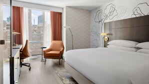 Een hotelkamer met een groot bed, een bureau, een bureaustoel, een staande lamp en uitzicht op de stad door grote ramen.
