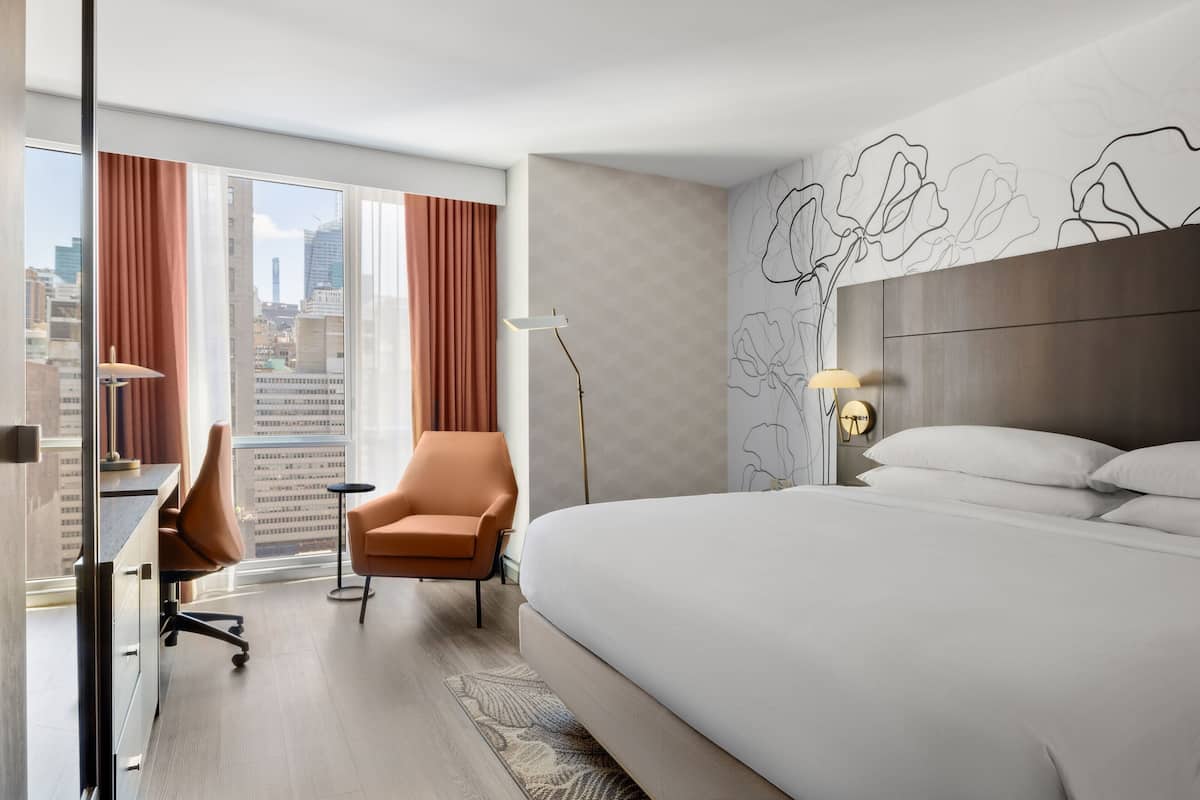 Ein Hotelzimmer mit einem großen Bett, einem Schreibtisch, einem Bürostuhl, einer Stehlampe und Blick auf die Stadt durch große Fenster.