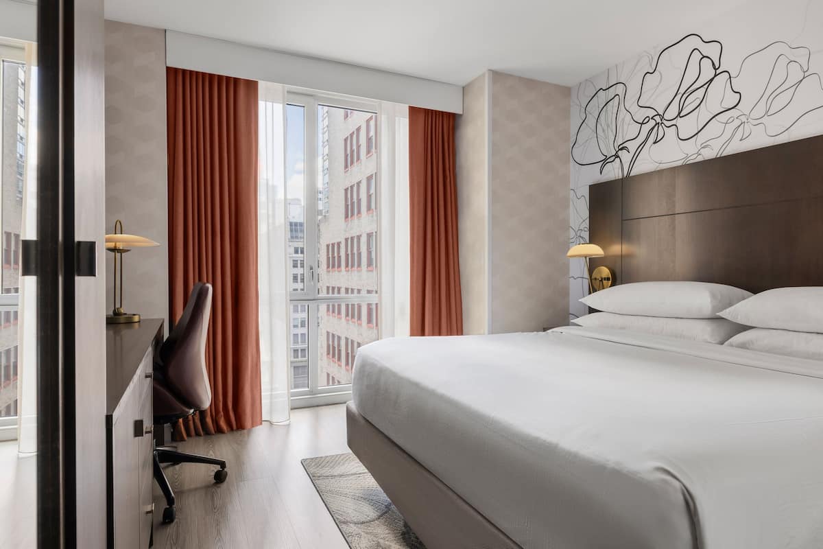 Ein Hotelzimmer mit einem großen Bett, einem Schreibtisch mit Stuhl, einer Lampe und Blick auf die Stadt durch das Fenster.