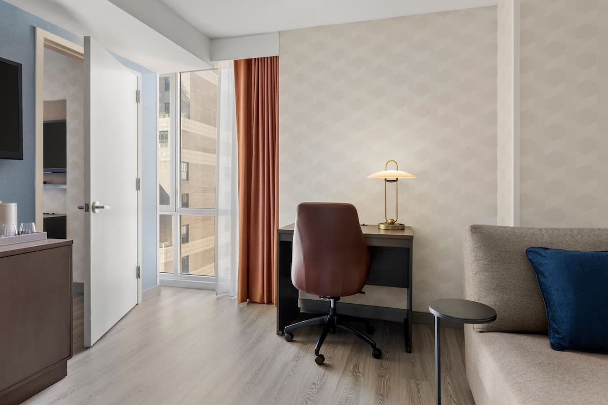 Ein Hotelzimmer mit Schreibtisch, Stuhl und einer Couch. Ein Fernseher ist an der Wand, eine Lampe auf dem Schreibtisch und ein Fenster mit Vorhängen.