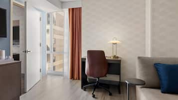 Ein Hotelzimmer mit Schreibtisch, Stuhl und einer Couch. Ein Fernseher ist an der Wand, eine Lampe auf dem Schreibtisch und ein Fenster mit Vorhängen.