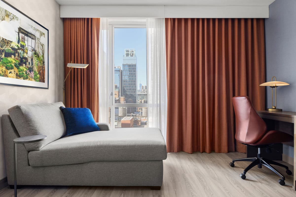 Ein modernes Hotelzimmer mit grauem Sofa, einem einzelnen Sessel, einem Schreibtisch und einem großen Fenster mit Vorhängen.