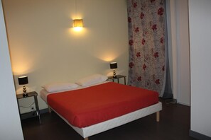 Habitación de hotel con cama, dos mesitas de noche, luz empotrada en la pared y cortina con estampado floral.