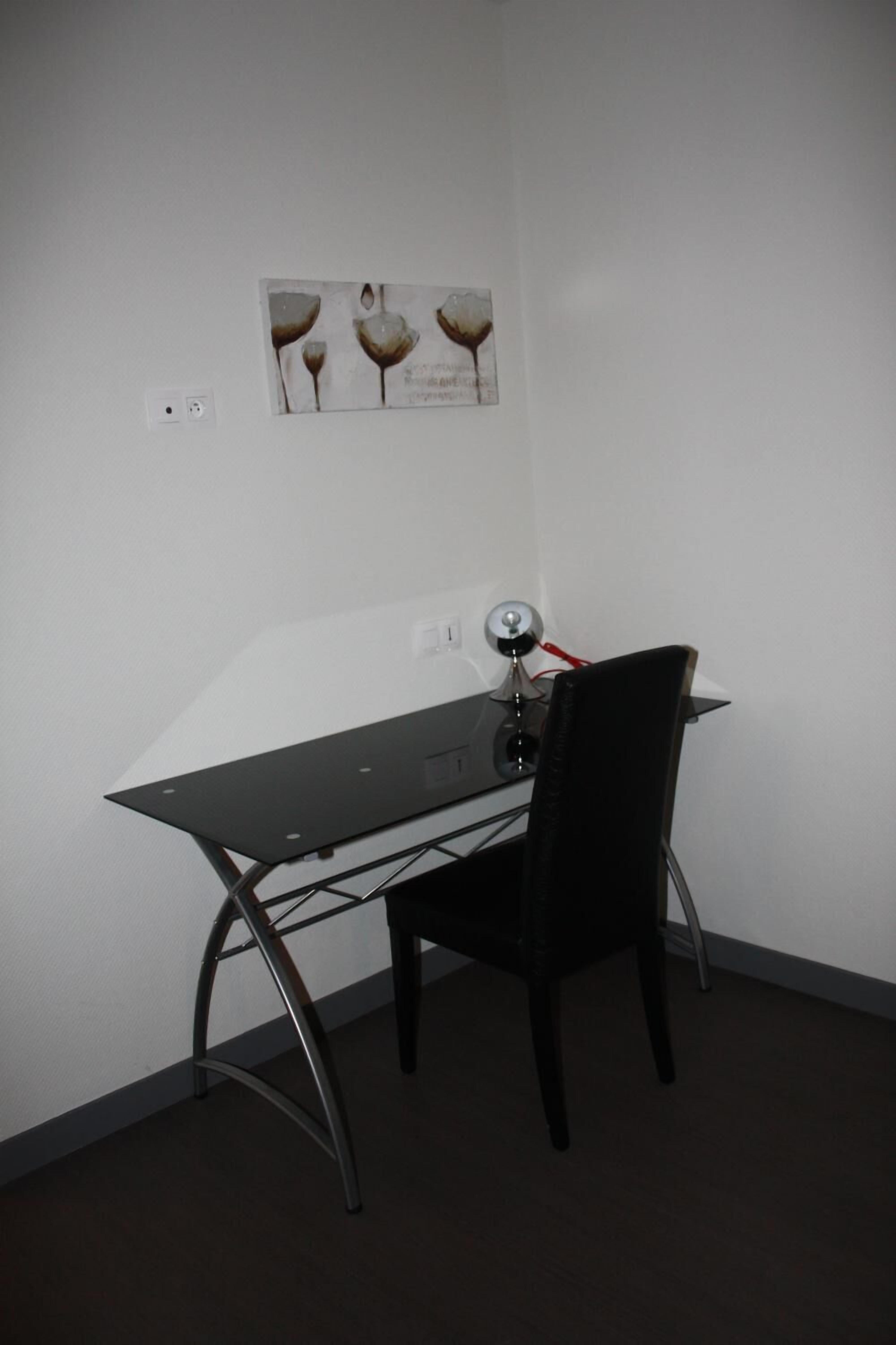 A small, dark-colored desk with a chair, a wall-mounted lamp, and a framed artwork above the desk.
