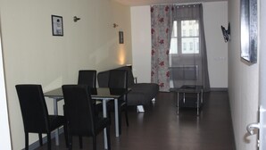 A dining area with a table and chairs, a sofa, and a window with floral curtains. - Résidence d'Artagnan (Montargis)
