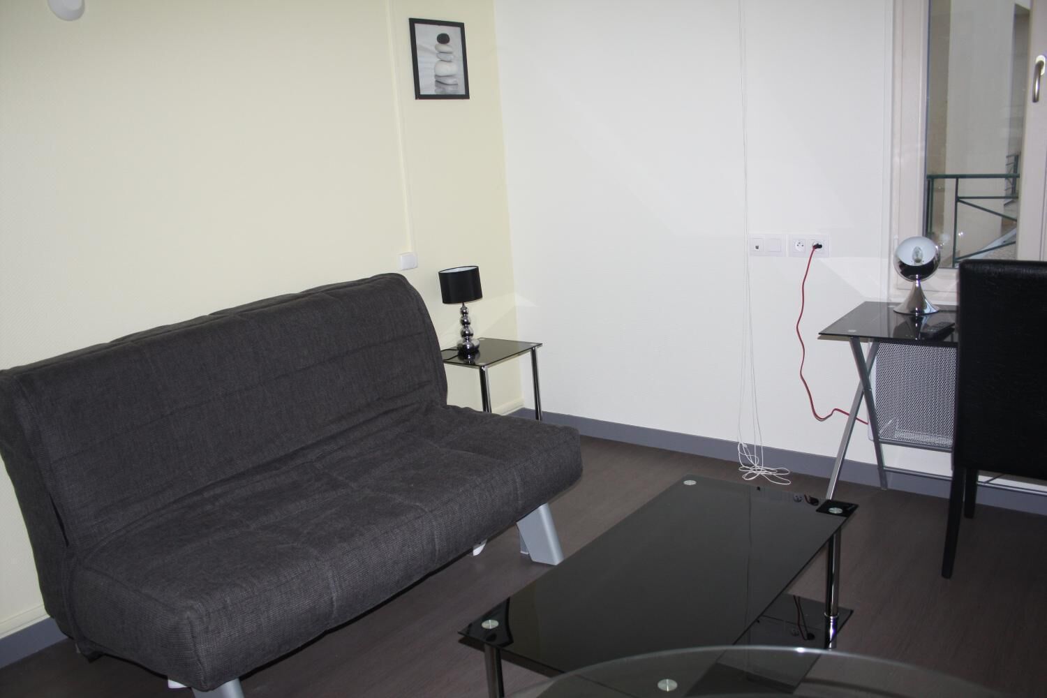 A modern living room with a grey sofa, a small black table with a lamp, and a glass coffee table.