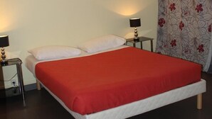 A bed with a red bedspread, two bedside tables with lamps, a wall-mounted light, and a floral-patterned curtain. - Résidence d'Artagnan (Montargis)