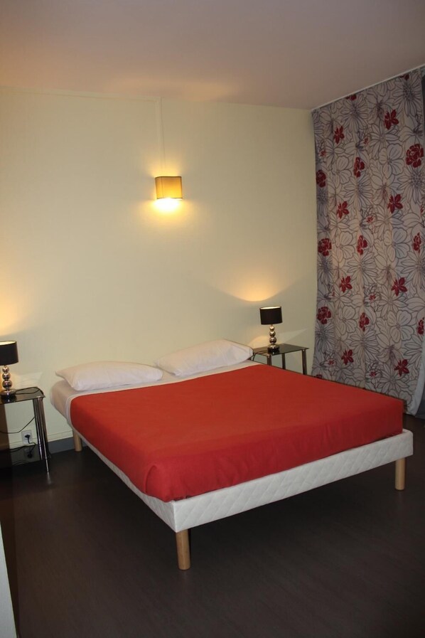 Una cama con colcha roja, dos mesitas de noche con lámparas, una luz empotrada en la pared y una cortina estampada con motivos florales.