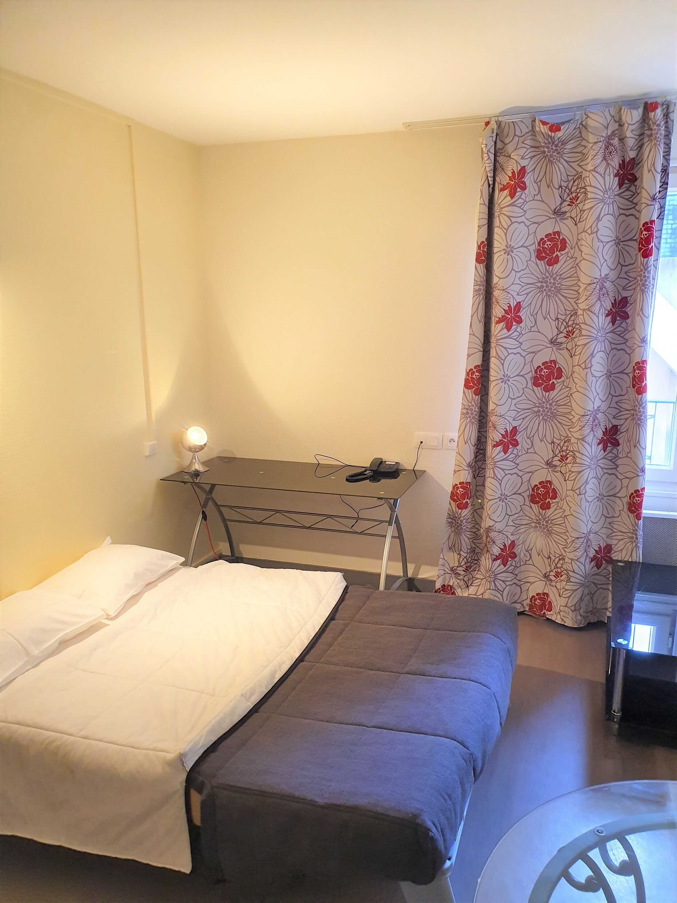 A small, simple room with a bed, a desk, and a chair. There is a window with a curtain.