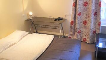 A small, simple room with a bed, a desk, and a chair. There is a window with a curtain.