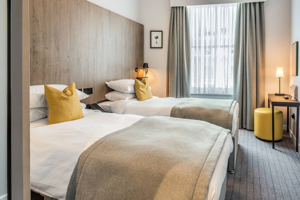A hotel room with two beds, a wooden headboard, a window with curtains, a bedside table with a lamp, and a small stool.