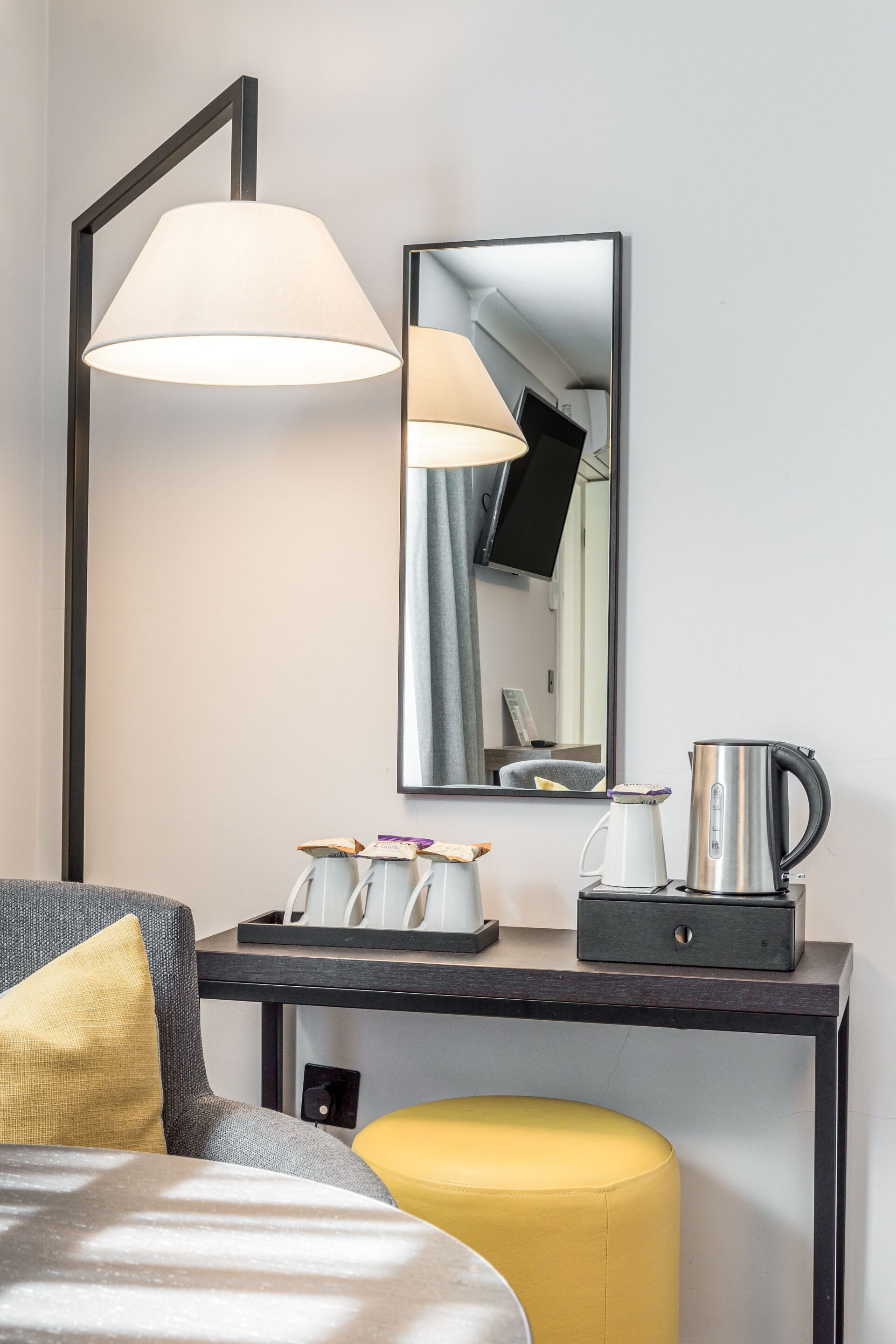 A hotel room with a bed, a desk with a lamp, a mirror, a coffee maker, and a small table with cups and a tray.