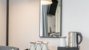 A hotel room with a bed, a desk with a lamp, a mirror, a coffee maker, and a small table with cups and a tray.
