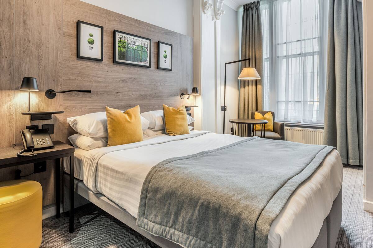 A hotel room with a large bed, two bedside tables with lamps, a small table with a chair, and a view of the outdoors through a window.
