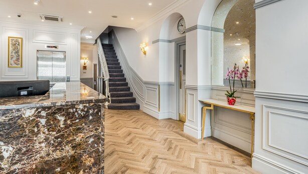 A hotel lobby with a marble reception desk, a staircase, and a reception desk with a vase of flowers. - The Princes Square Hotel (London)
