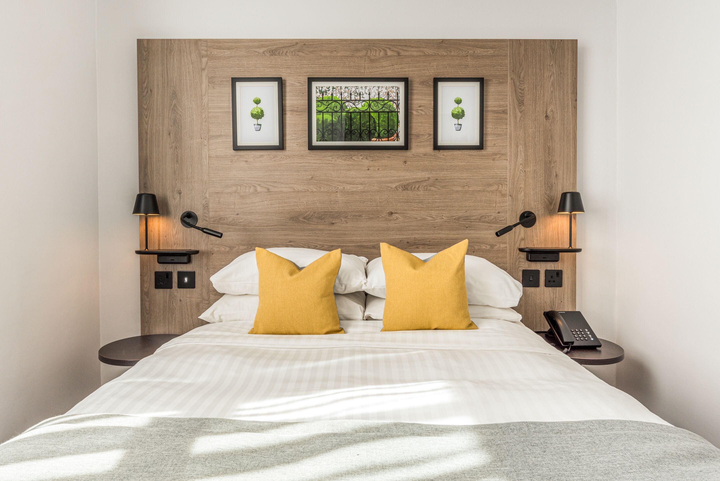 Cama bem arrumada com roupa de cama branca e duas almofadas amarelo-mostarda, com duas mesas de cabeceira laterais com abajures pretos e um telefone. Acima da cama, um painel de cabeceira de madeira com três quadros emoldurados.