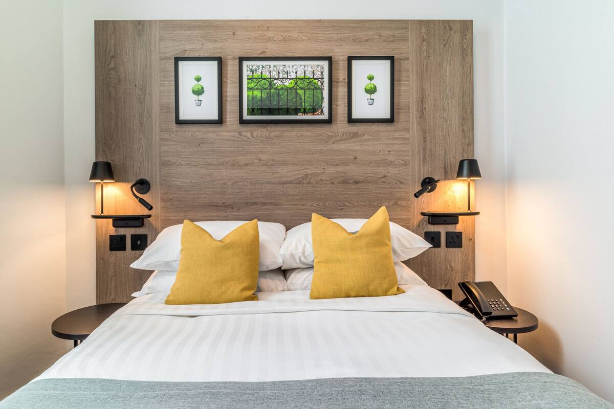 A hotel room with a wooden headboard, a bed with white bedding and two mustard pillows, two bedside lamps, a framed picture of a fence with greenery, and a telephone on a bedside table.