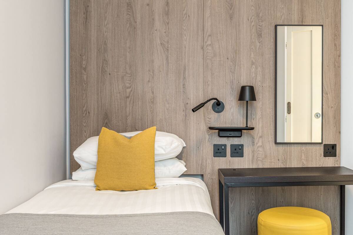 A hotel room with a bed, a nightstand, a lamp, a mirror, and a wooden wall.