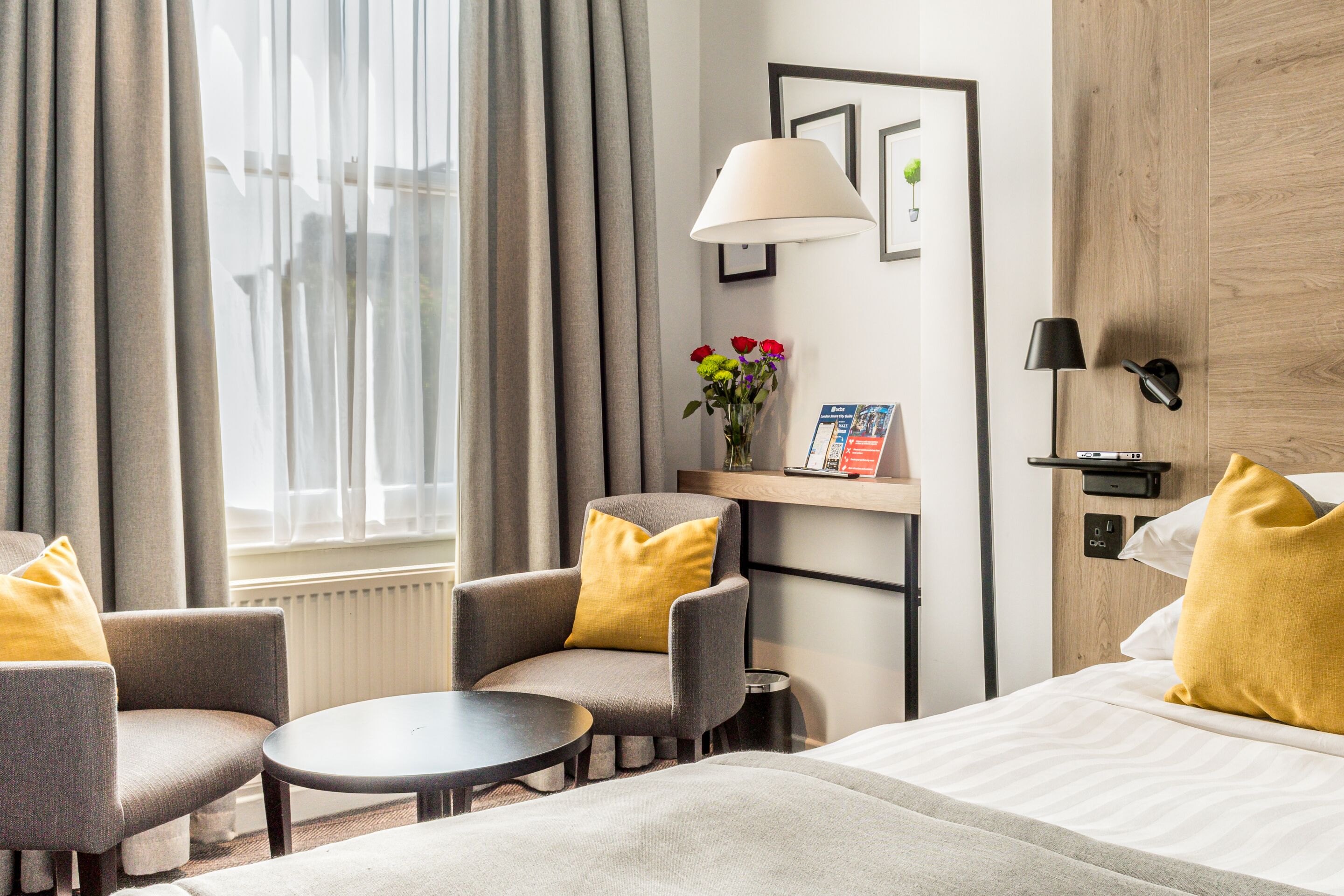 Ein Hotelzimmer mit einem Bett, zwei Sesseln, einem kleinen Tisch, einer Lampe, einer Vase mit Blumen und einem Regal mit Buch und Pflanze.